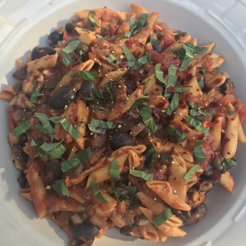 a serving bowl of penne pasta puttenesca topped with strips of basil