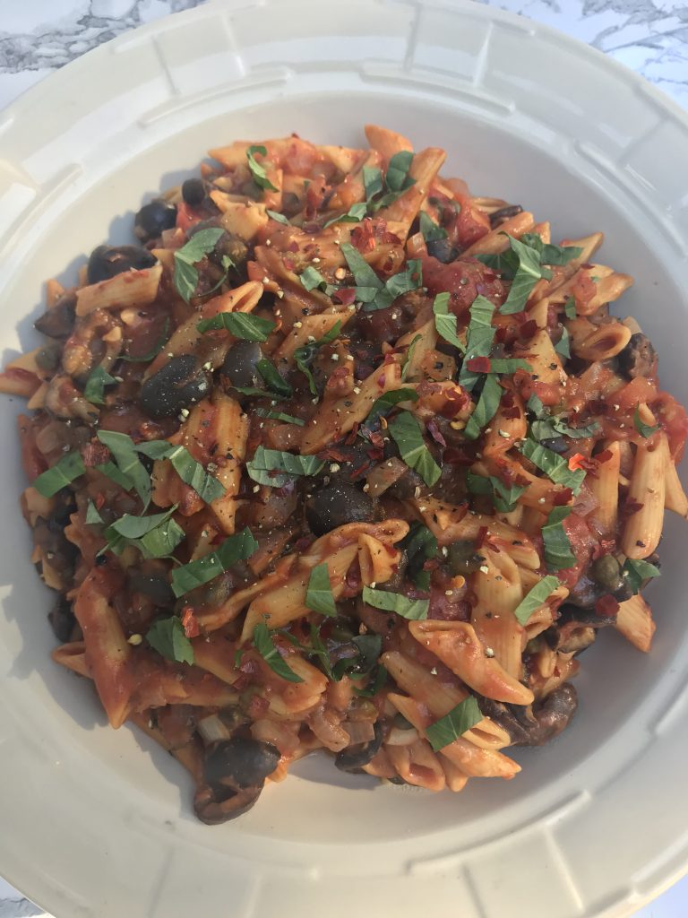 a serving bowl of penne pasta puttenesca topped with strips of basil