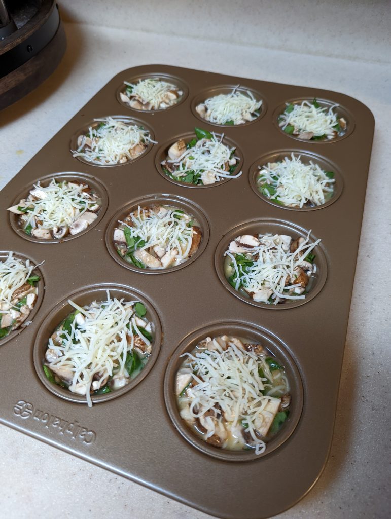 Uncooked spinach and mushroom breakfast muffins with cheese on top in a muffin tin