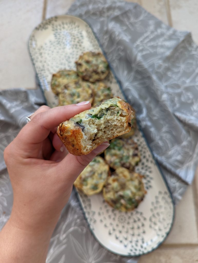 A white hand holding a muffin with a bite taken out. The platter of the rest of the muffins is in the background.