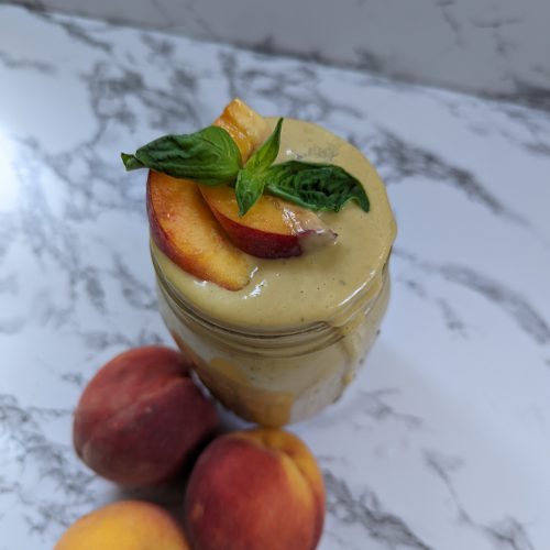 A glass jar filled with creamy peach basil smoothie with a peach wedge and basil leaf garnish