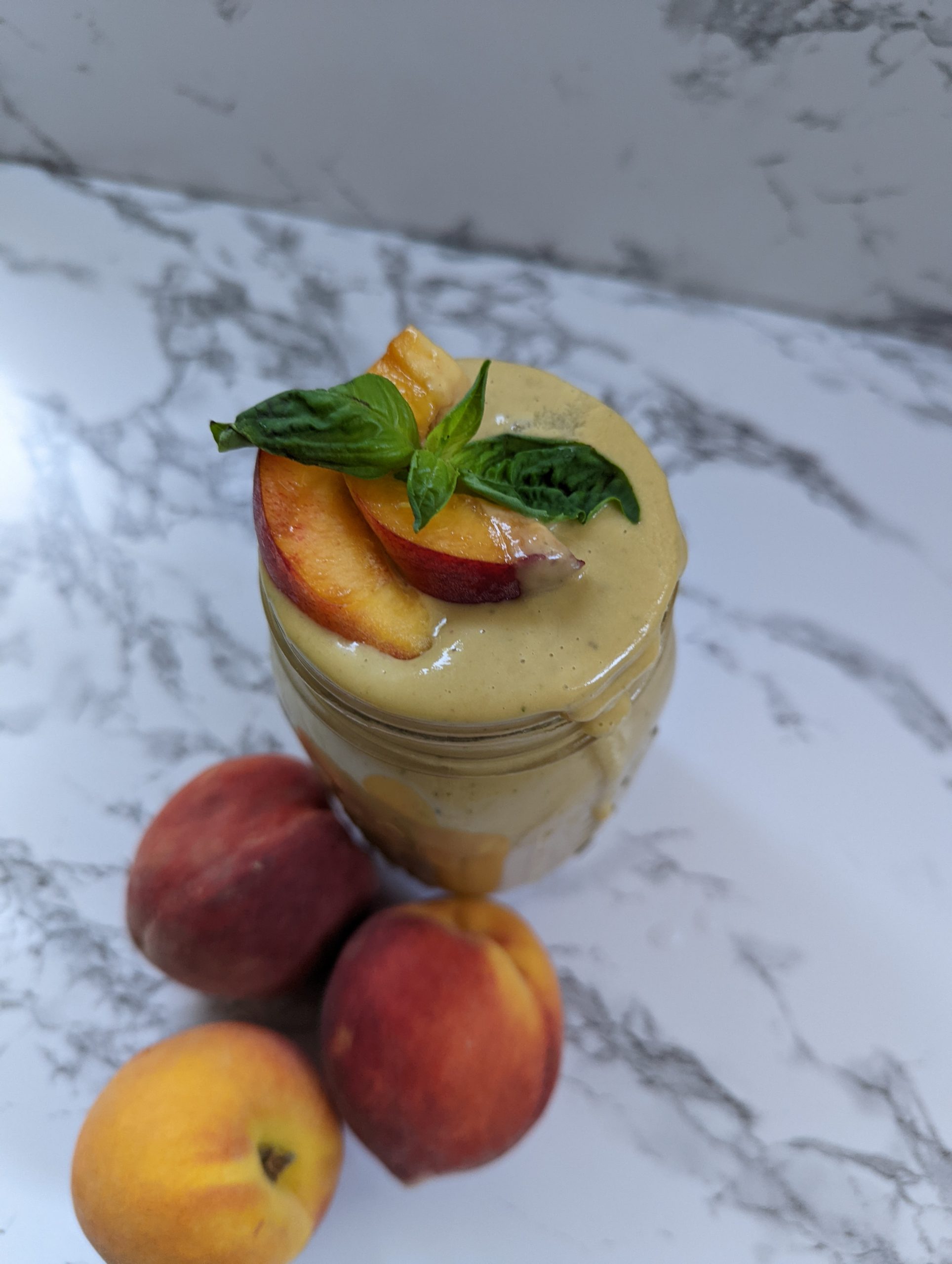 A glass jar filled with creamy peach basil smoothie with a peach wedge and basil leaf garnish