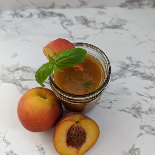 a mason jar filled with a peach margarita with a basil garnish. There is a whole peach and a half or a peach with the pit exposed around the glass.