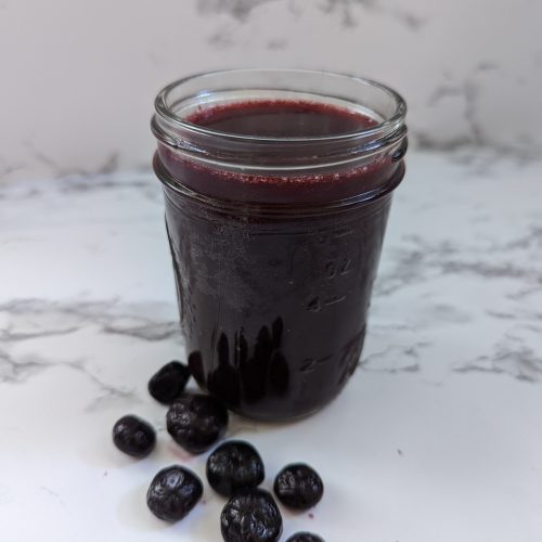 A mason jar filled with a deep purple blueberry margarita with some blueberries next to the glass.